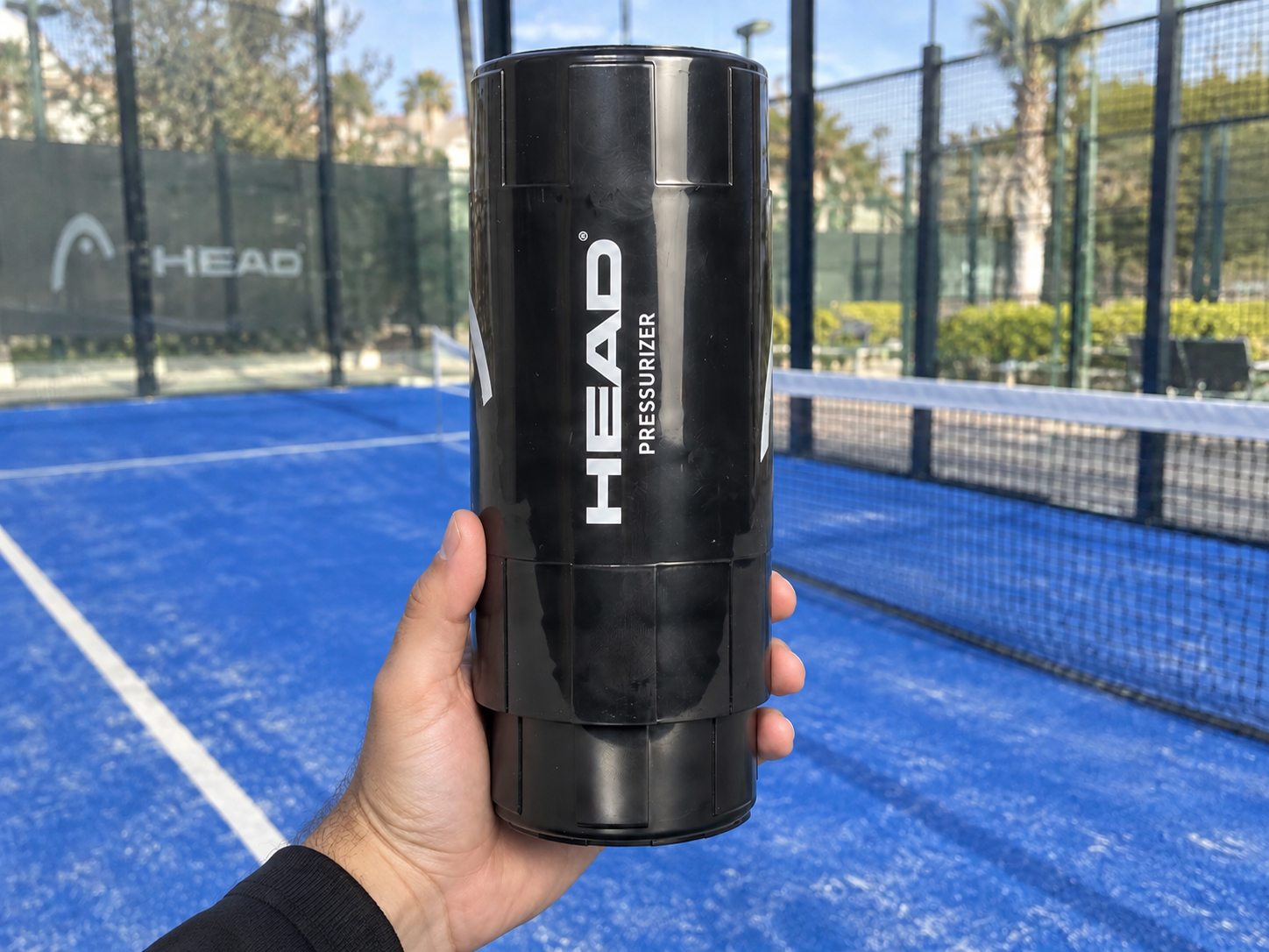 Presurizador de pelotas de pádel HEAD sostenido en la mano junto a una pista de pádel azul